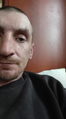 The video shows a close-up of a man's face. He has short, thinning hair and a goatee. He is wearing a black sweatshirt. The background is a reddish-brown wooden surface and a white pillow. The man looks directly at the camera with a serious expression.
