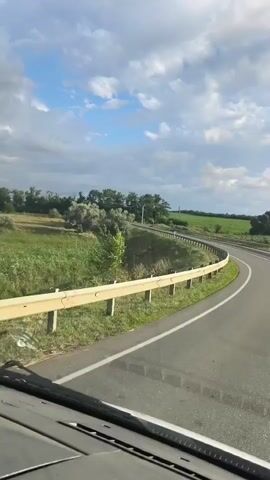 The video shows a view from the front of a vehicle driving on a paved road that curves to the right. There is a white metal guardrail on the left side of the road, separating it from a grassy area with some bushes and trees. The sky is partly cloudy with patches of blue sky visible. The road has a white line marking the center and the edge. The interior of the vehicle, including the dashboard and windshield wipers, is visible in the foreground.