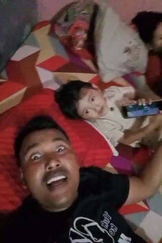 A man is taking a selfie video on a bed. He is in the foreground, with his mouth open in a wide smile. Behind him, a child is lying on a red pillow, holding a smartphone. The child appears to be looking at the phone. To the right of the child, a woman is lying on the bed, also looking at a smartphone. The bed has a patterned comforter. The lighting suggests an indoor setting, possibly a bedroom. The overall mood of the video appears to be lighthearted and familial.