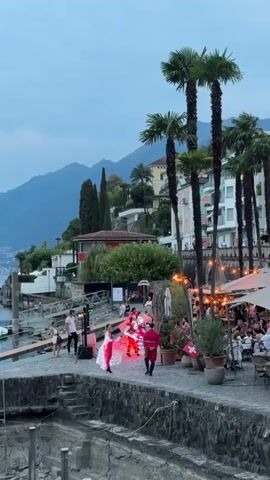This video captures an outdoor evening scene with a body of water and mountains in the background. Palm trees line a paved walkway where several people are visible. In the foreground, a few individuals are near a DJ setup. Center frame, two performers are engaged in an activity. One, dressed in a red outfit, dances while the other, in a luminous white outfit, moves in sync. A group of people are seated at tables in an outdoor dining area to the right, some with umbrellas for shade. The lighting suggests dusk, with artificial lights illuminating the performance area and string lights overhead at the dining establishment.
