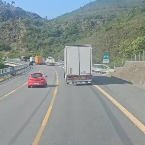 The video shows a view from a vehicle traveling on a highway behind a white semi-trailer truck. The truck has "LeciTrailer" written on the back and speed limit indicators of 90, 80, and 70. A small red car is visible in the left lane. The highway is bordered by a metal guardrail and a steep, vegetated hillside. The sky is clear and blue.