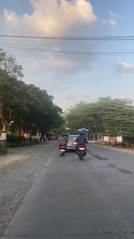 This is a vertical, eye-level, outdoor, daytime video taken from the perspective of someone riding a vehicle on a paved road. The road has a grey surface with some cracks and patches. On the left side of the road, there is a gravel shoulder with some vegetation and small buildings. On the right side of the road, there are more trees and some buildings. The sky is visible with a few clouds and appears to be late afternoon due to the lighting.

In the foreground, there are two motorcycles riding in the same direction as the camera. The motorcycle on the left is closer to the camera and has a rider wearing a dark jacket and a helmet. The motorcycle on the right is slightly further away and has a rider wearing a dark jacket and a helmet. Both motorcycles are riding on the paved road.

In the mid-ground, there is a silver car driving in the same direction as the motorcycles. Further back, a blue and white truck is visible. There are also other vehicles and people sparsely scattered in the distance. Trees line both sides of the road, creating a somewhat shaded environment in places. The overall scene depicts a typical street scene with traffic on a sunny day.