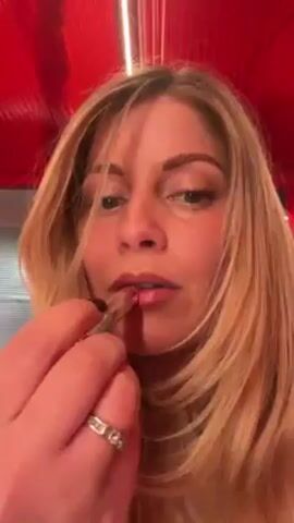 A woman is applying lipstick in a close-up shot. She has long, blonde hair and is wearing a ring on her finger. The lipstick tube is silver and the color is a shiny rose. The background appears to be a red ceiling with a light fixture and a glimpse of a store. She is looking directly at the camera while applying the lipstick.