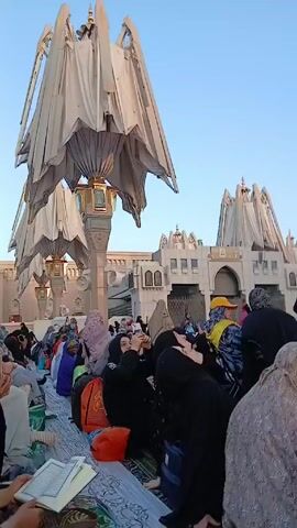 The video shows a crowd of people gathered in what appears to be an outdoor area near a building with large, open, decorative structures that resemble umbrellas. Many of the individuals are sitting on the ground, some are taking pictures with their phones, and others are looking around. Most of the people appear to be wearing traditional Islamic clothing. The environment looks peaceful, with clear blue skies in the background.