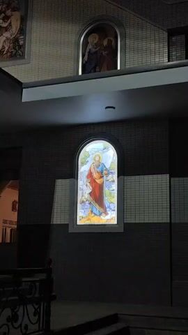 The video shows a portion of a building's exterior at night. The building appears to be a church or a similar religious institution, as evidenced by the stained-glass windows depicting religious figures and scenes. The windows are illuminated from within, casting a warm glow on the facade. The architecture features arched windows set into tiled walls, with a covered entrance area supported by columns. The scene is lit by artificial lights highlighting the architectural details and the stained-glass art. The overall impression is one of solemnity and quiet beauty in the evening light.