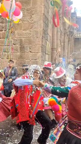 People are dancing in a street celebration. They are wearing brightly colored clothing and hats, and some are covered in white foam. Balloons and streamers are visible in the background. The celebration is taking place in front of a stone building. Some people are taking photos with their phones. The atmosphere appears festive and lively.