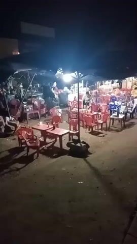 The video shows an outdoor seating area next to a street at night. The seating area consists of numerous plastic tables and chairs under a dark canopy, illuminated by overhead lights. Some tables have bottles on them. The ground is dirt. A person is briefly visible under the canopy. The camera moves from the seating area towards the street. Cars are parked along the sidewalk, and other vehicles, including a truck, are driving on the street. A sign with illuminated text is visible across the street.