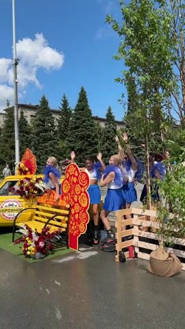 A group of young women in blue and white outfits, some wearing large blue mesh collars, are gathered around a yellow car decorated with flowers. One woman is on roller skates. They are in an outdoor setting, possibly a street or a park, with trees and buildings in the background under a blue sky with clouds. The women appear to be posing or performing, with some raising their arms and smiling. A decorative element with red and gold floral patterns is also visible. The scene seems festive and celebratory.