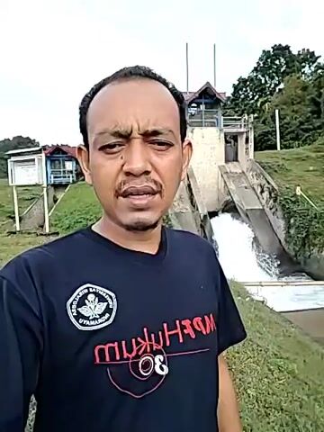 A man is standing outside on a grassy area, with a dam in the background. He is wearing a black t-shirt with writing on it. The dam has a small building at the top and water is rushing down its concrete slope into the water below. There are trees and another building in the background as well. The man looks directly at the camera. At the start of the video he is blowing smoke.