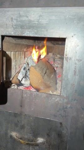 A close-up shot shows a metal wood-burning stove with a fire burning inside. The fire is burning on a large chunk of wood, with flames reaching upward. There is ash and embers around the wood. The interior of the stove is lined with brick. The stove door is ajar, revealing the fire inside. There is some conversation in the background.
