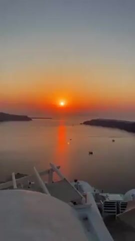 The video shows a sunset over the sea, likely from Santorini, Greece, given the white-washed architecture in the foreground. The sun is bright orange and casts a strong reflection on the water, where several small boats are visible. In the distance, land masses frame the seascape. The sky transitions from a lighter orange near the sun to a pale grey at the top of the frame.