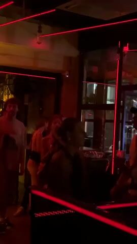 A group of people are gathered indoors, possibly a bar or club, illuminated by red lights. In the foreground, a person wearing a black top is seated. Behind them, a line of people are walking. To the left, a man in a grey t-shirt and shorts is visible, as well as another man with glasses. The background shows a bar area with bottles on shelves. Red laser lights are strung across the ceiling.