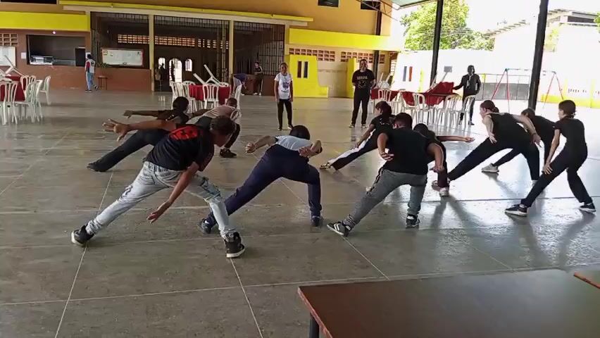 The video shows a group of around 10 people in a large, open-air, covered space doing a dance or exercise routine. They are spread out, all facing forward, with their legs wide apart, arms extended to the sides, and leaning from side to side. Most of the people are wearing casual clothing. An individual, likely an instructor, stands in the middle observing and directing the group. In the background, there are tables with red tablecloths, chairs, and other individuals standing or walking around. The setting appears to be a community center or recreational hall. The space has a light green metal roof structure and yellow and beige colored walls with rectangular window openings.
