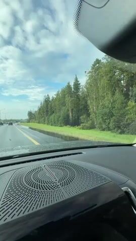 The video is filmed from the perspective of a passenger or driver inside a vehicle, looking forward through the windshield. The vehicle is on a multi-lane highway with other cars in the distance. The road appears wet, possibly from recent rain. To the right of the highway is a grassy verge bordered by a dense forest of green trees, with some birch trees visible. The sky is cloudy with patches of blue. In the foreground, the dashboard of the vehicle is visible, including a large speaker grille with a circular pattern. The audio is of a song or voice, possibly Russian, with lyrics about walking and starting something.