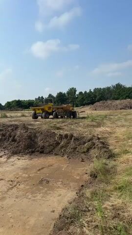 The video shows an outdoor construction site on a sunny day with a blue sky and scattered white clouds. In the foreground, there is uneven dirt ground with some grass and piles of soil or debris. In the mid-ground, a large yellow dump truck is visible, parked on a dirt surface. Further in the background, there are trees, some buildings, and what appears to be more piles of dirt or compost. The camera pans slowly across the scene.