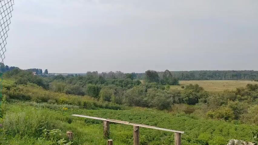 The video shows a wide, panoramic view of a rural landscape. In the foreground, a green, grassy slope is visible, with some wooden posts and a plank forming a simple railing or barrier. Beyond this, a vast expanse of green vegetation, likely fields and trees, stretches into the distance. Several trees and bushes are scattered throughout the middle ground, with a winding body of water, possibly a river or stream, partially visible. In the far distance, a treeline forms a dark band against the pale sky, and a few scattered buildings and structures can be seen on the horizon to the left. The sky is overcast and a hazy, muted color. The overall impression is one of a calm, natural, and undeveloped environment. The camera appears to be stationary, capturing a static, wide shot of the scenery. There are no people or animals visible in the frame. The lighting suggests it is daytime, likely late morning or afternoon given the brightness.