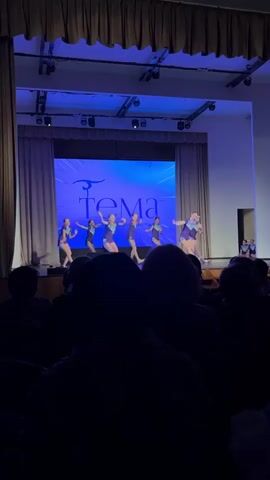 A group of young dancers performs on a stage with blue lighting in front of an audience. The stage has a screen in the back with the words "Тема клуб современного танца" displayed on it. The dancers are wearing matching purple costumes and are executing a choreographed routine. The audience is largely silhouetted, with only the backs of their heads visible. The stage is flanked by draped curtains and overhead stage lights.