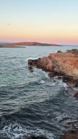 This is a vertical, medium shot of a rocky coastline at dusk. The sun is setting, casting warm hues of pink and orange across the sky and illuminating the reddish-brown rocks that jut into the turquoise sea. Waves crash against the shore, creating white foam that contrasts with the dark, choppy water. In the distance, a low, sandy island is visible, shrouded in the soft light of the fading day. The scene is tranquil and serene, with no people or man-made objects present.