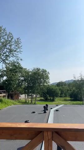 A video pans across a wooden deck overlooking a paved area with some black cylindrical objects and a metal grate. Beyond the paved area, there are trees, grass, and a body of water with a distant treeline. The sky is clear blue with a few wispy clouds. The camera movement is smooth and steady.