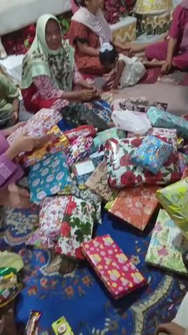 The video shows a group of people, predominantly women and some children, gathered indoors. They are sitting on the floor, which is covered with a patterned rug. In front of them is a large pile of wrapped gifts, many of which are brightly colored and decorated with floral patterns. Some of the women are wearing hijabs. In the background, there is a sofa, some decorative items, and what appears to be a large, ornate basket. The lighting suggests it is daytime or a well-lit indoor space. The overall impression is a festive gathering, possibly a celebration or gift-giving event.