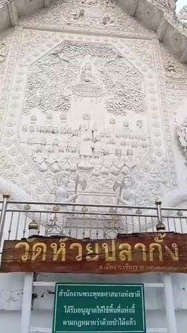 A low-angle, medium shot shows the facade of a white temple with intricate relief carvings. The carvings depict a central figure beneath a tree, surrounded by numerous smaller figures. Below the carvings, there is a metal railing and two wooden signs. The top sign has golden Thai lettering, and beneath it is a smaller green sign with Thai text. The sky above is overcast.