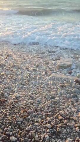 The video opens on a pebbled beach with waves gently washing ashore. The sun, a bright orb, is setting over the ocean, casting a warm golden glow across the water. As the video progresses, the sun sinks lower, and the sky turns a soft orange. An airplane can be seen flying high in the clear blue sky. The waves continue to roll in, their white foam contrasting with the darkening sea. The scene is peaceful and serene, capturing the beauty of a coastal sunset.