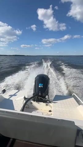 This media file shows a view from the back of a speeding motorboat on a body of water, likely a lake or calm sea. The camera is positioned low, looking forward over the stern of the boat. Dominating the foreground is the boat's outboard motor, a black unit with a blue emblem that reads "60 EFI". The motor is churning the water behind the boat, creating a prominent white wake that stretches back towards the horizon.

The water is dark blue and choppy from the boat's movement, reflecting the bright sky. The sky itself is a vibrant blue, with scattered white, fluffy clouds that suggest fair weather. The sun is visible on the left side of the frame, casting a bright glare on the water and creating lens flare.

In the distance, a thin line of dark green foliage marks the shoreline, indicating land. The overall impression is one of speed, freedom, and enjoyment on a sunny day. The scene suggests a recreational activity, likely a leisurely boat trip or watersports. The bright sunlight and blue sky convey a sense of warmth and pleasantness. The location is identified as Siukterka, Russia.