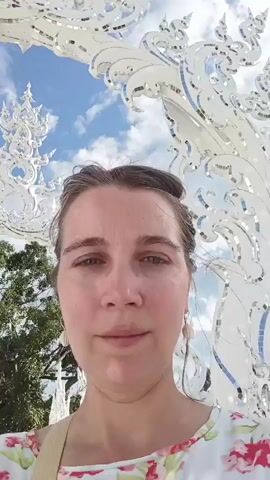 The video shows a medium close-up of a woman's face. She has brown hair pulled back, light skin, and is wearing a white shirt with a floral pattern. She is looking directly at the camera with a neutral expression. Behind her, there is a large, ornate white structure that appears to be made of glass or mirrored material, with intricate swirling designs. The structure is illuminated by sunlight, and reflects the bright blue sky with white clouds. In the background, there are trees and more of the white structure. The overall impression is of an outdoor setting, possibly a tourist attraction or a place of worship. The video is shaky, suggesting it was filmed by someone holding the camera themselves.