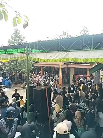 This video shows an outdoor gathering of people. In the foreground, there are several large speakers, suggesting some form of performance or event. A crowd of people, many wearing headscarves, are gathered in front of and around the speakers, watching something happening to their left. Further back, there is a building with an open-air, covered walkway. More people are visible in and around this building. The overall atmosphere appears to be lively, with a sense of community engagement.