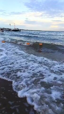 The video shows a view of the ocean from a beach. The water is blue and there are waves crashing on the shore. The sky is cloudy and there are some boats in the distance. There are also some orange buoys in the water. The camera is positioned low, near the water's edge, and is pointed towards the ocean. The overall impression is one of a peaceful beach scene.