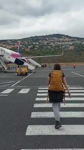A woman with a brown backpack is walking on a crosswalk toward an airplane on an airport tarmac. The airplane is white and pink with a set of stairs connected to its door. Several people are at the base of the stairs and on the tarmac. In the background, a hillside is populated with houses. The sky is overcast.