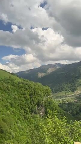 The video pans across a lush green valley with mountains in the background. The sky is partly cloudy with patches of blue. A small village with red-roofed buildings is nestled in the valley. In the foreground, a tree with sparse branches is visible, with other green foliage and a wooden fence. The overall impression is one of natural beauty and tranquility.