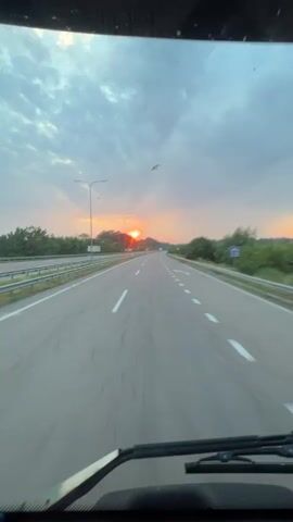 The video shows a view from inside a vehicle driving on a highway at sunrise. The sun is visible on the horizon, casting a warm glow and creating a dramatic sky with clouds. The highway has multiple lanes and is bordered by trees and vegetation. The motion blur indicates the vehicle is moving at a moderate speed.
