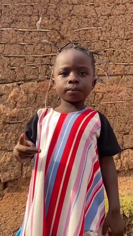 This is a medium shot video of a young Black child, likely a toddler, standing in front of a rustic mud-brick wall. The child has dark skin, dark eyes, and short, dark hair styled with twigs or small branches sticking up, creating a crown-like effect. The child is wearing a colorful dress with a black neckline and sleeves, and the main body of the dress features bold vertical stripes in red, white, light pink, and light blue. The child's left hand is raised, with the index finger pointing slightly forward. The background is a textured brown mud wall, suggesting an outdoor setting, possibly in a rural or traditional environment. The lighting appears to be natural daylight, casting some shadows and highlighting the child's face. The child's expression is neutral, perhaps a bit serious. The video is filmed at a slightly low angle, looking up at the child. The setting, Ufu Esioni, Nigeria, provides a geographical context for the scene, hinting at a West African cultural backdrop. There is no visible text within the frame. The overall impression is a portrait of a child with a unique hairstyle, set against a simple, natural background.