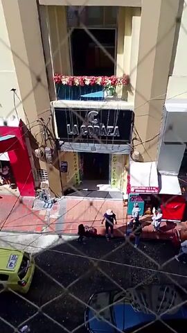 From an elevated perspective behind a wire fence, the video shows a street scene with several businesses including "La Coraza", "Piko Riko", "UKEY", "Cid Express", and "Calzados SK".  People are walking and milling about on the sidewalks. There is a yellow taxi parked near orange traffic cones. Cars and motorcycles move along the street. One person is pushing a cart. Some individuals are carrying boxes or bags. There's general pedestrian activity with people engaged in what appears to be shopping or other errands.