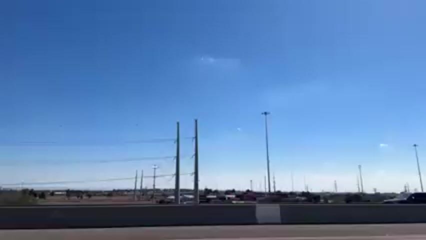 The video shows a view from a moving vehicle on a highway. The sky is blue and mostly clear. On the side of the highway, there are power lines, light poles, and some buildings in the distance. A white SUV passes on the opposite side of the highway.