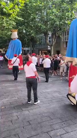 A street procession features several very large, costumed figures that appear to be giants. Two of these giants are prominently visible, one on the left wearing a blue robe and another on the right, also in a blue robe with red trim and a golden headdress. People are walking behind and around the giants, many dressed in casual clothing. Some individuals are wearing red berets and white shirts, suggesting a uniform for participants in the procession. One person in the foreground is playing a drum. The procession is taking place on a paved street lined with buildings that have balconies and windows. The time of day appears to be late afternoon or early evening, with the sky visible at the top of the frame. The overall atmosphere seems festive.