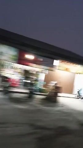 The video shows a street scene at dusk or night. There are several small shops or food stalls lining the street, some with lights on. A motorcycle is parked in front of one of the buildings. In the distance, headlights of vehicles can be seen on the road, suggesting traffic. The overall atmosphere is dimly lit and appears to be a commercial or residential street.