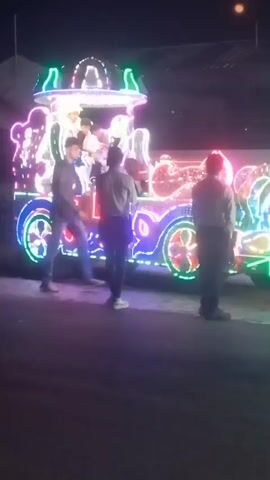 At night, a decorated vehicle with people aboard is parked on a street. The vehicle is brightly lit with green, red, blue, and white lights and has a dome-like structure on top.  A group of people stand around the vehicle. A building is visible in the background.