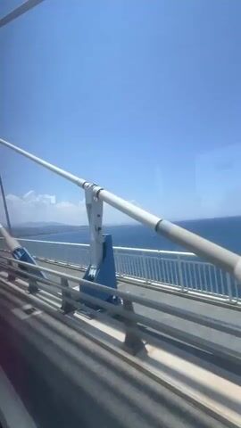 The video shows a view from a moving vehicle, likely a bus or car, on a bridge. The camera is positioned to capture the scene through a window. The dominant colors are the bright blue of the sky and the deep blue of the ocean. A white railing runs along the side of the bridge, separating the roadway from the water. In the foreground, parts of the bridge's structure are visible, including large white and blue metal components. A long white pipe-like structure, possibly part of the bridge's suspension system, extends diagonally across the frame. The ocean stretches to the horizon, and a distant coastline with faint clouds can be seen. The overall impression is one of travel over water on a clear, sunny day.