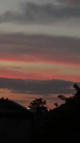 The video shows a sky at sunset. There are horizontal stripes of pink and orange clouds across the sky. The bottom of the frame is dark, showing the silhouettes of rooftops and trees.