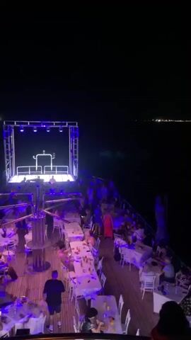 The video captures a night scene on a boat, likely a party or dining cruise. The camera pans across a deck filled with people seated at white-clothed tables, suggesting a meal service or social gathering. To the left, a brightly lit stage or DJ booth with blue and red lights is visible, hinting at entertainment. The background shows a dark expanse of water with distant city lights visible along the horizon. The atmosphere appears lively, with many individuals present and engaged in conversation or enjoying the ambiance.