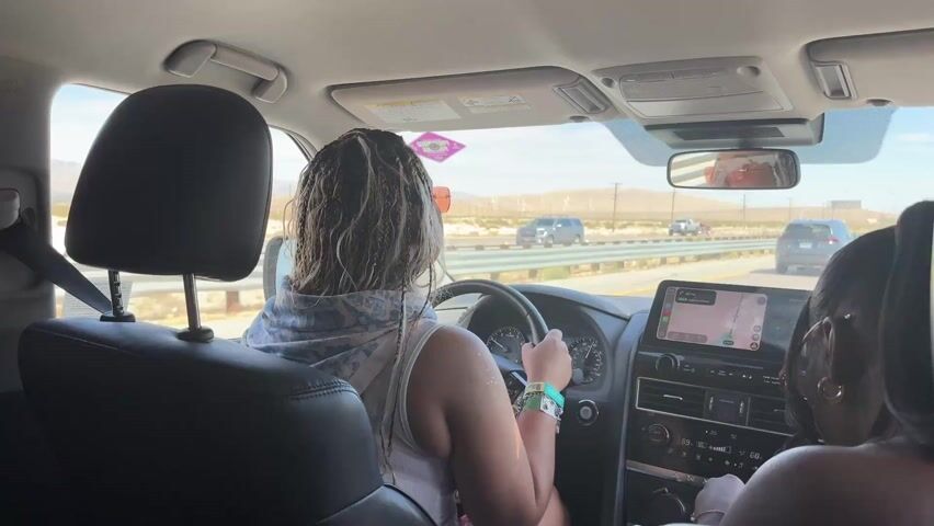 The video shows the interior of a car driving down a highway on a sunny day. The driver, a woman with braided hair, is visible from the back seat, wearing sunglasses and a white tank top. Another person is in the passenger seat. A navigation system is displayed on the car's infotainment screen. Wind turbines are visible in the distance, indicating a desert environment. The camera perspective is from the back seat, focusing on the driver and the road ahead.
