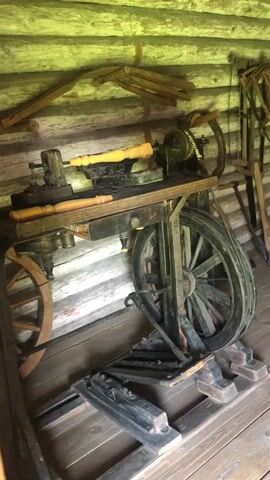The video shows the interior of a wooden log cabin. In the foreground, there is a large, antique woodworking lathe with a heavy iron base and a large wooden wheel. Various wooden tools and pieces of wood are visible on and around the lathe. In the background, shelves are mounted on the log walls, displaying numerous wooden buckets and other historical artifacts. The overall impression is of a historical workshop or storage space.