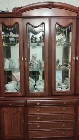 The video shows a tall, dark wooden display cabinet filled with a collection of white porcelain dishes and silver-colored serving pieces. The cabinet has glass doors on its upper section, revealing multiple shelves. The items on display include teacups, saucers, plates, pitchers, and decorative bowls. Some of the silver pieces appear to be part of a tea set. In the reflections on the glass doors, a person can be seen standing and observing the cabinet's contents. The cabinet is situated in what appears to be a domestic interior, likely a dining room or living room, given its size and the nature of its contents. The lighting suggests it's daytime, and there are no obvious indications of specific weather conditions. The overall scene conveys a sense of quiet admiration or appraisal of a well-maintained collection. There is no visible text in the video.