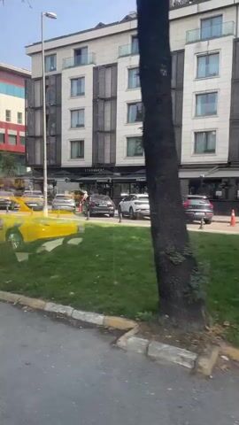 The video shows a street scene with a Starbucks on the ground floor of a modern building. Cars are parked along the street and in front of the Starbucks. There are trees and grass in the foreground. The video is taken from a moving vehicle, likely a car or bus, looking out the window. The scene appears to be in a city, with several buildings visible in the background. The weather appears to be sunny and clear.