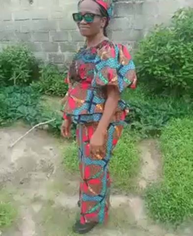 A woman stands outdoors, wearing a vibrant, colorful patterned top and skirt that match. She also has a matching head wrap and dark sunglasses. The outfit features a mix of red, turquoise, brown, and white patterns. She stands on a dirt path surrounded by patches of grass and small bushes. Behind her is a cinder block wall. The lighting is natural daylight. The woman is smiling slightly, and the camera seems to be focused on her.