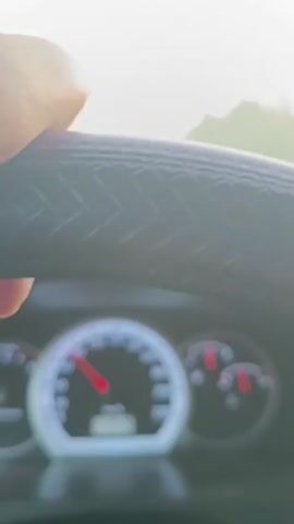 The video shows a first-person perspective from inside a car driving down a road. The driver's side dashboard and steering wheel are visible in the foreground. The speedometer shows the car is traveling at approximately 40-50 km/h. The road is paved and has lane markings. Trees and foliage line both sides of the road, forming a tunnel-like effect. The sky is overcast and appears to be either early morning or late evening, with a soft, diffused light. There are no other vehicles or people visible on the road. The overall impression is one of a quiet, scenic drive.