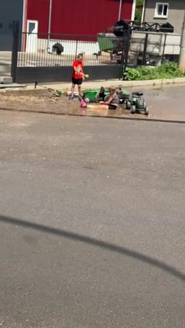 A young boy in a red shirt and dark shorts is playing with a green toy tractor and a doll on a wet patch of asphalt, possibly from a sprinkler. Another child, a girl in a dark top and pink shorts, is lying on her stomach near the tractor, also interacting with the toys. The scene appears to be a residential street, with a building with a red facade and a white upper story in the background. There is also a black metal fence and some vegetation visible. The video captures a moment of outdoor play.