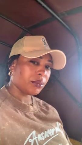 The video shows a woman in a beige Ralph Lauren baseball cap and a brown t-shirt with the word "Answer" printed on it riding in a yellow and maroon tricycle vehicle. The tricycle is moving on a city street with buildings visible in the background. The driver of the tricycle, a young man wearing a white shirt and carrying a black backpack, is seen briefly in the middle of the video. The woman looks directly at the camera and occasionally smiles.  Sunlight enters the tricycle from the front.